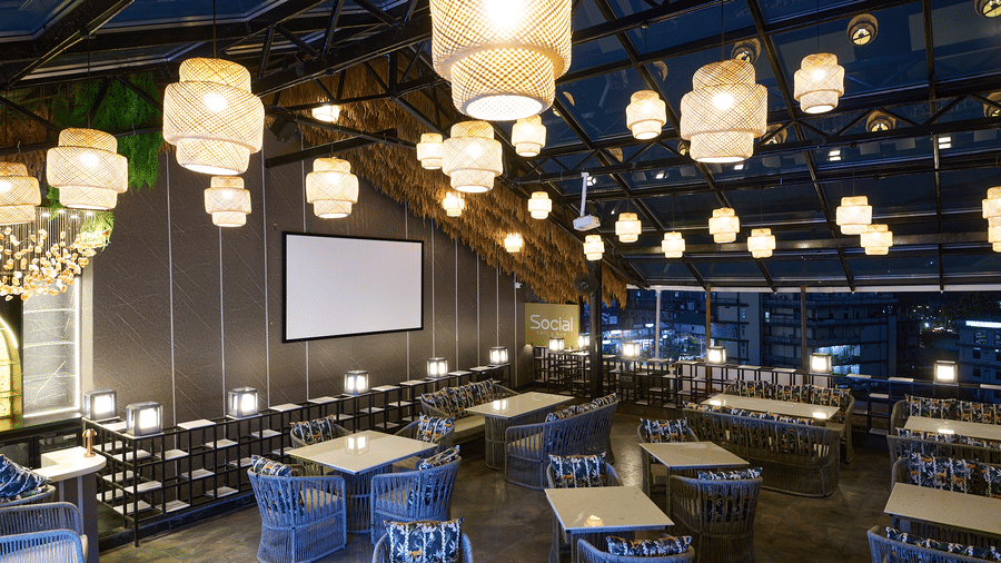 An indoor restaurant or dining area with blue chairs and tables, illuminated by multiple hanging lantern-style lights, and a projection screen in the background | Voyage Eastin Suites