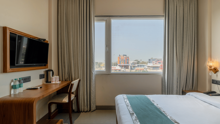 Twin bedroom at Hotel Highway Prince, Behror, with two beds, a wall-mounted TV, a long wooden desk, and a window with grey curtains.