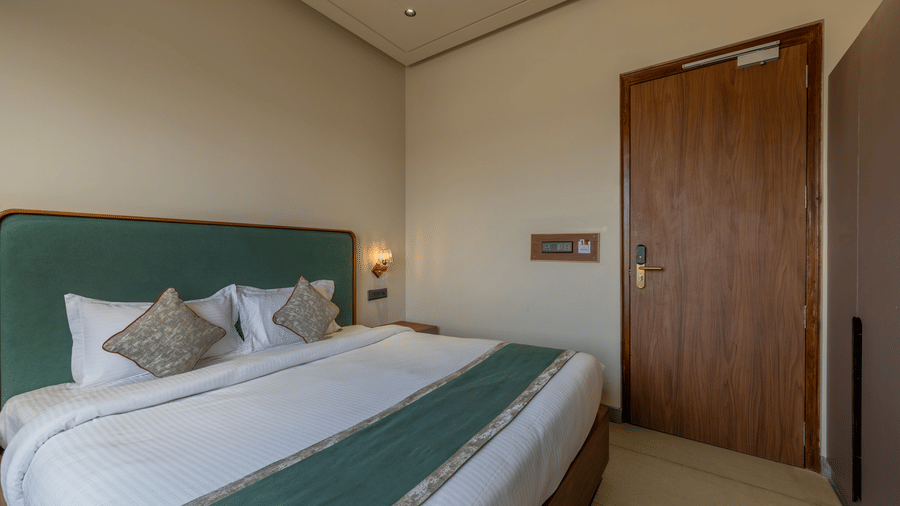View of a room at Hotel Highway Prince, Behror, showing a large bed with a teal headboard and runner, a wooden door, and a wall-mounted light switch panel.