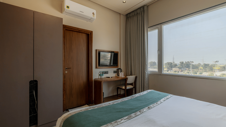 A plush double bed in a Deluxe Room at Hotel Highway Prince, Behror, facing a wall-mounted TV placed in a corner of the room with a work desk right below, and a large glass window beside it with the curtains drawn.