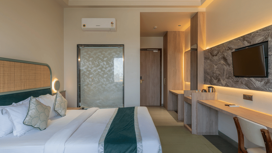 Spacious layout of Super Deluxe Room showcasing the plush double bed with pillows and cushions facing the wall-mounted TV placed on a panel, an a frosted glass bathroom door.