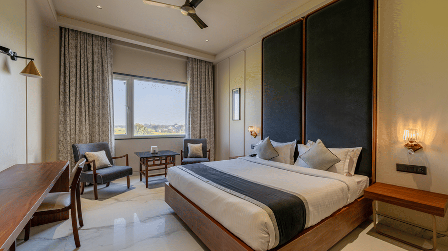 Bedroom at Hotel Highway Prince, Behror, featuring a king bed with a green runner, a wooden desk, a ceiling fan, and a large window.