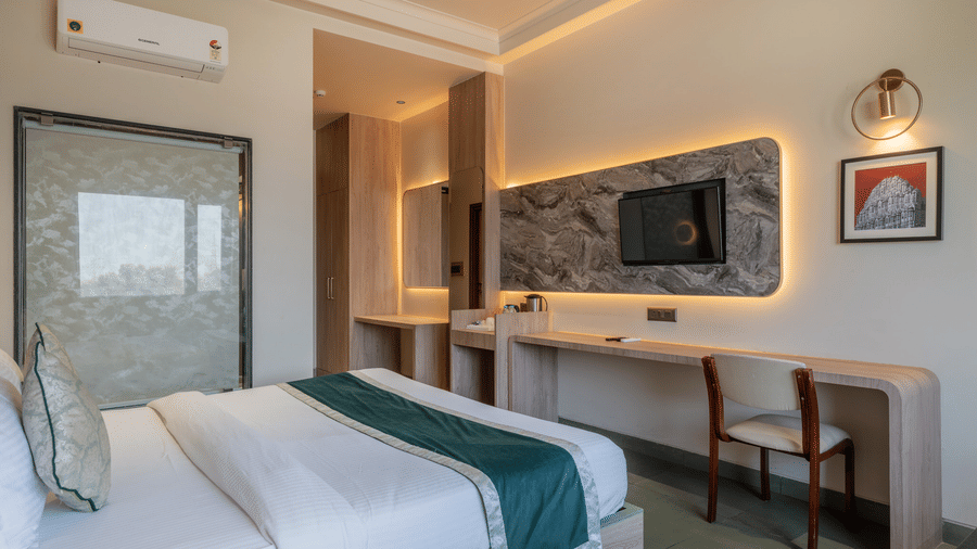 Modern bedroom at Hotel Highway Prince, Behror featuring a large bed, wall-mounted TV, a long wooden desk with a chair, and warm recessed lighting.