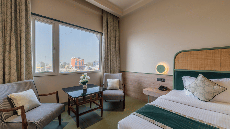  Perspective view of a Hotel Highway Prince, Behror guest room with a double bed, a sofa by the window, and a wooden side table.