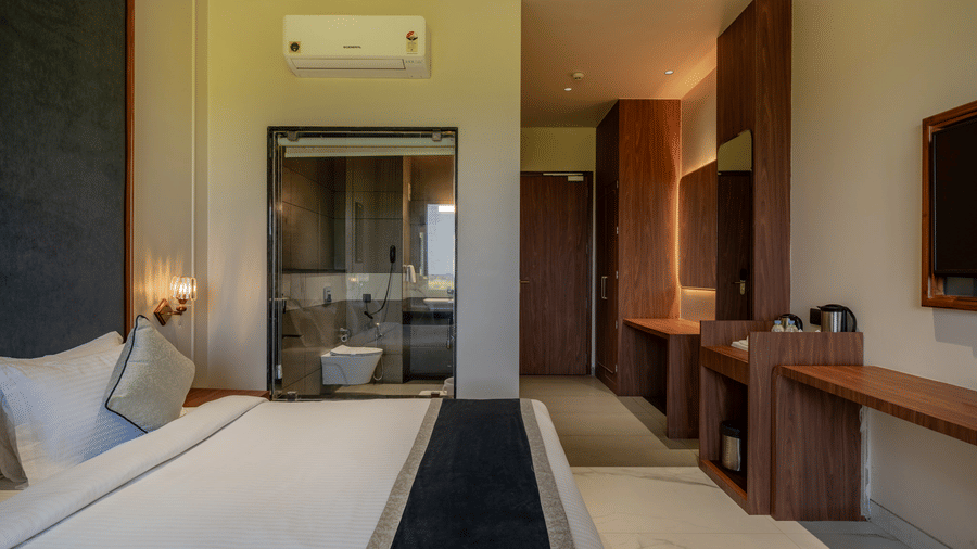 A sleek bedroom view at Hotel Highway Prince, Behror, showing a large bed, wall-mounted air conditioner, and a hallway leading to the bathroom.
