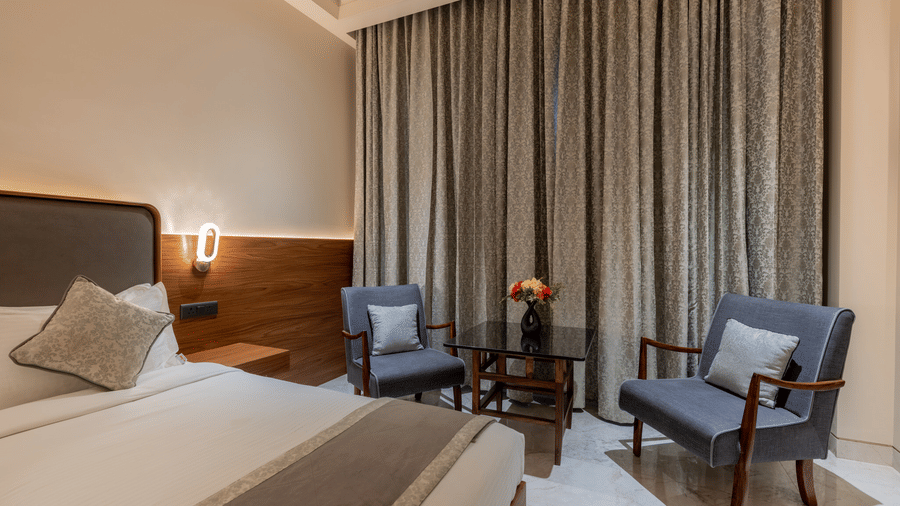Modern bedroom interior at Hotel Highway Prince, Behror, with a large bed, wall-mounted headboard, armchairs, and heavy curtains over the window.