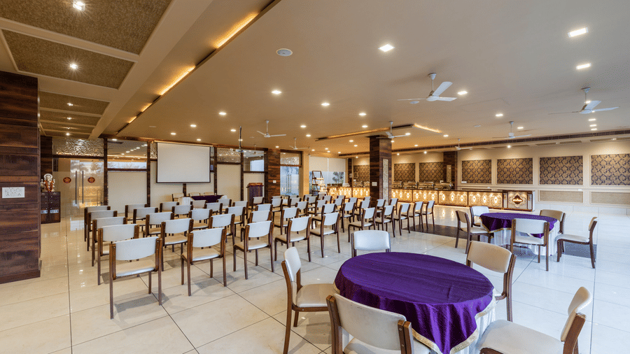 Hotel Highway Prince Paota wide-angle banquet hall view with white and wooden chairs, purple table covers, projector screen, and warm ceiling lights.