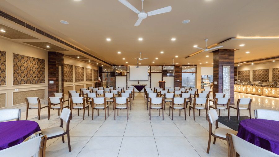 Hotel Highway Prince Paota banquet hall with neatly arranged wooden chairs, purple-draped tables, decorative wall panels, and ceiling fans.