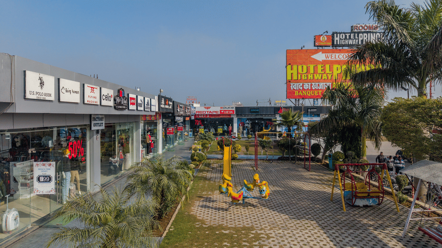 Hotel Highway Prince Paota aerial forecourt view with paved entrance, palm trees, colourful signboards, parked vehicles, and retail outlets.