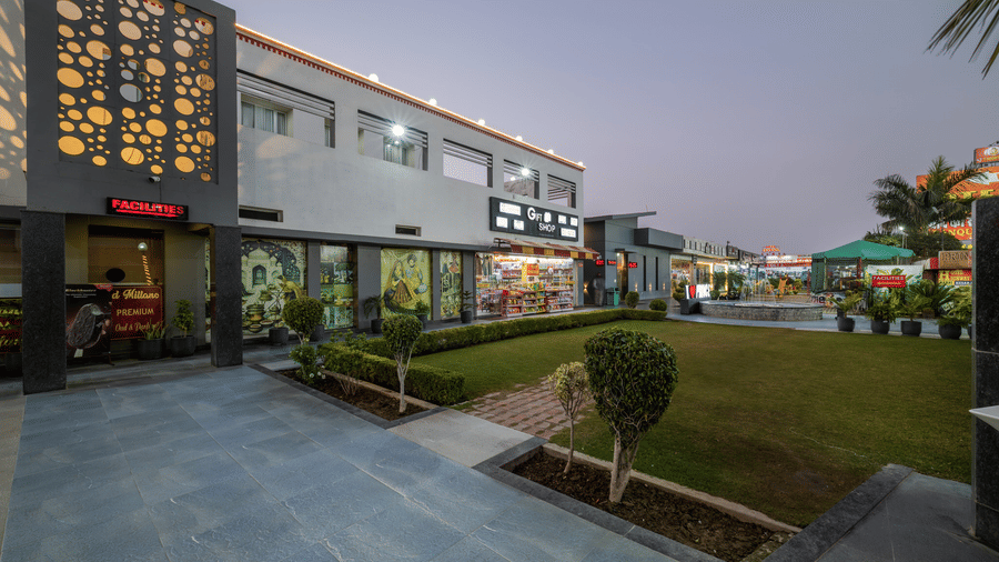 Hotel Highway Prince Paota exterior dusk view with decorative perforated facade, illuminated retail outlets, manicured shrubs, and twilight sky.