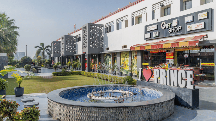 Hotel Highway Prince Paota morning exterior with circular fountain, I Love Prince signage, retail shops, green lawn, and bright daylight.