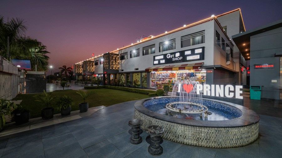 Hotel Highway Prince Paota circular fountain with I Love Prince branding at dusk, surrounded by lit retail outlets and twilight purple sky.