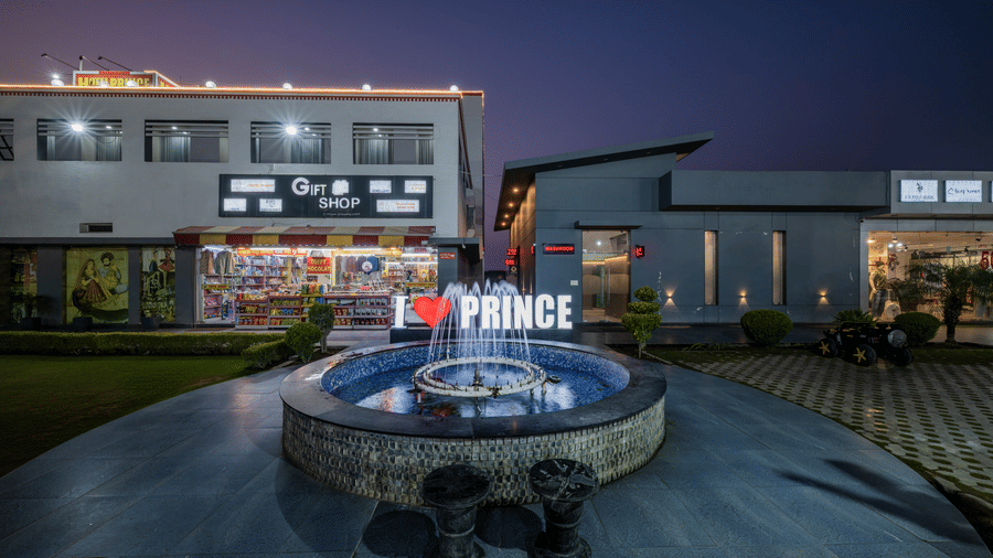 Hotel Highway Prince Paota illuminated circular fountain at night with I Love Prince signage, lit retail shops, and vibrant evening atmosphere.