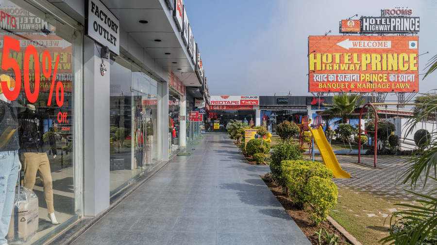 Hotel Highway Prince Paota highway-facing frontage with large billboards, retail shops, pedestrian walkway, potted plants, and colourful signage.