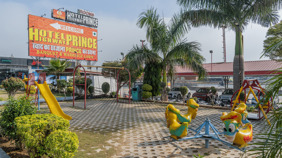 Hotel Highway Prince Paota highway-facing plaza with large illuminated hotel signboard, palm trees, colourful bird sculptures, and retail frontage.