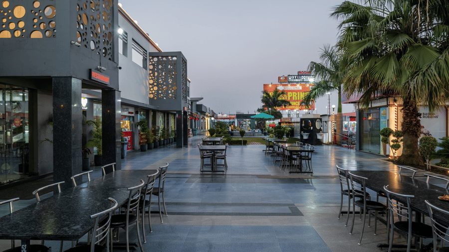 Hotel Highway Prince Paota daytime retail promenade with outdoor seating, paved walkway, palm trees, decorative facade, and clear sky.