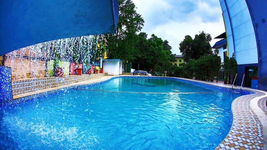 Assymetrical pool at Krushnai Resort with a partly cloudy sky, featuring an artificial waterfall created by a blue-coloured concrete slab on one side.