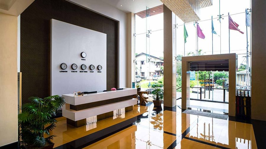 A hotel lobby at Mansha Hotel Regency featuring a white reception desk, reflective floors, and large windows with natural light overlooking the exterior with flags.
