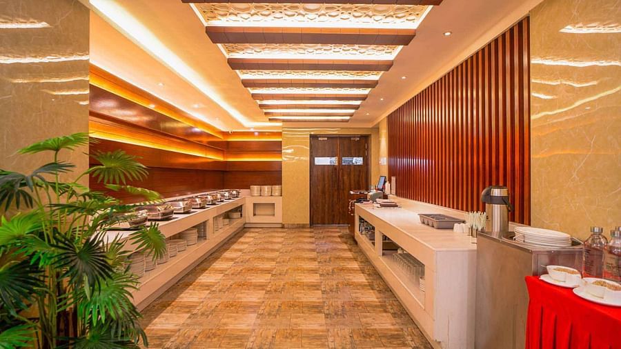 A bright, long hallway featuring a buffet counter with a red tablecloth and green potted plants, leading towards a dining area at Mansha Hotel Regency.