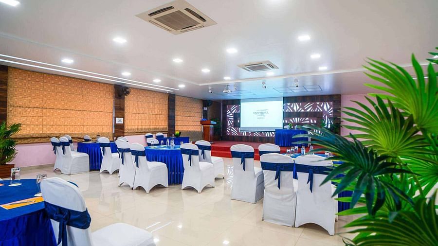 A brightly lit banquet hall at Mansha Hotel Regency with round tables draped in blue and white tablecloths, set with chairs, and green plants as decoration.