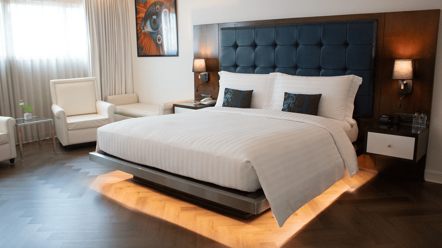 An overview of the Grand Gold King Room's bedroom with illuminated platform bed, tufted headboard, vibrant artwork, cream armchairs, and warm ambient lighting throughout at Night Hotel Bangkok in Sukhumvit - the best district to stay in Bangkok.