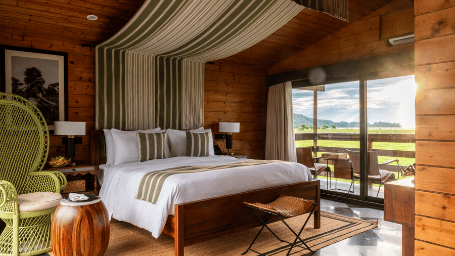 A luxury suite at Rhino & River Wildlife Retreat & Spa, Pobitora, featuring a wooden bed, a decorative striped ceiling canopy, and a green wicker chair.