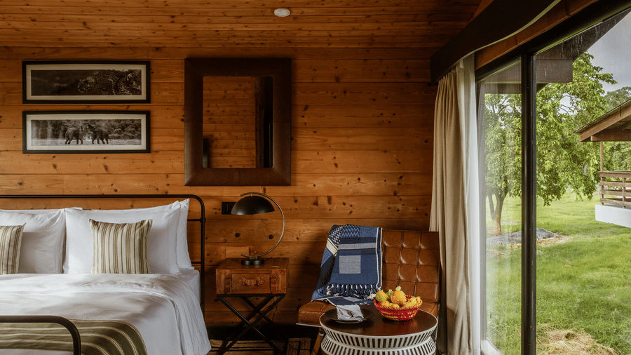 A view inside a wooden bedroom at Rhino & River Wildlife Retreat & Spa, Pobitora, showing a bed with white linens next to a floor-to-ceiling glass window.
