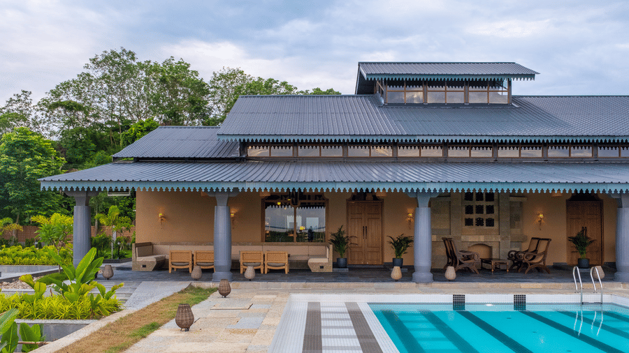 A wide exterior view of the main building at Rhino & River Wildlife Retreat & Spa, Pobitora, featuring traditional architecture, a stone patio, and a clear blue swimming pool under a cloudy sky.
