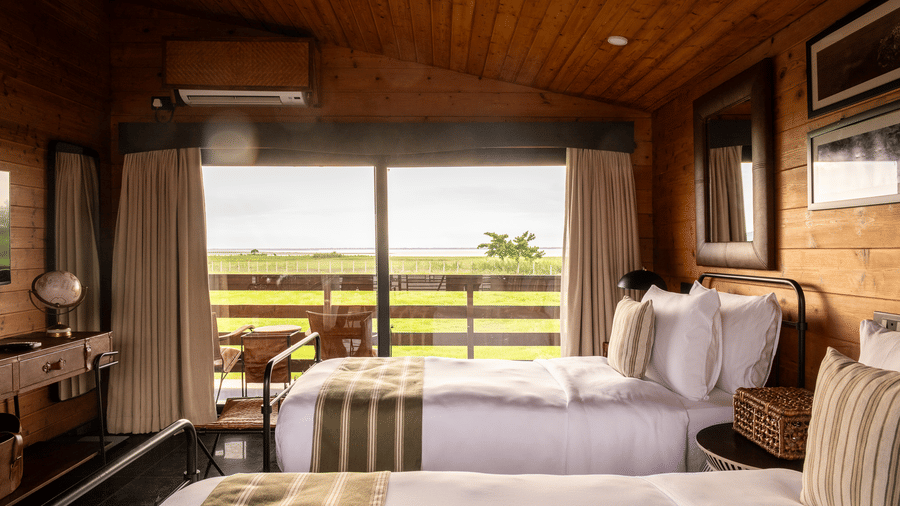A guest room at Rhino & River Wildlife Retreat & Spa, Pobitora, with two twin beds featuring black metal frames, set against a backdrop of large windows overlooking green fields.