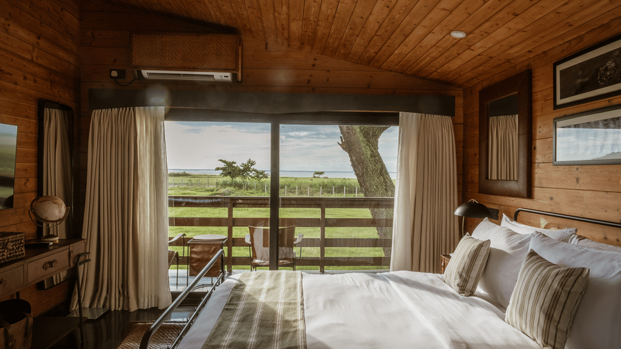 An interior shot of a bedroom at Rhino & River Wildlife Retreat & Spa, Pobitora, showing a bed with striped pillows and a wide view of the sunny plains through the window.