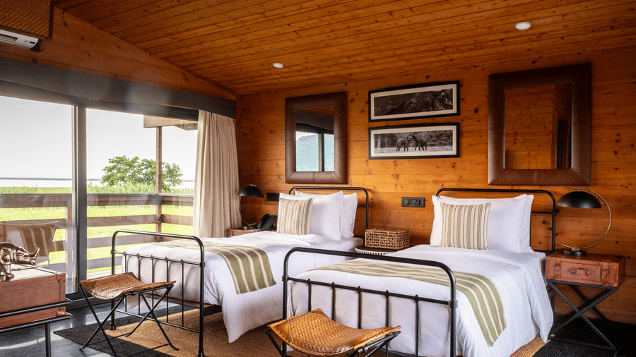A wide interior shot of a twin bedroom at Rhino & River Wildlife Retreat & Spa, Pobitora, showing the transition from the wooden sleeping area to the private balcony.