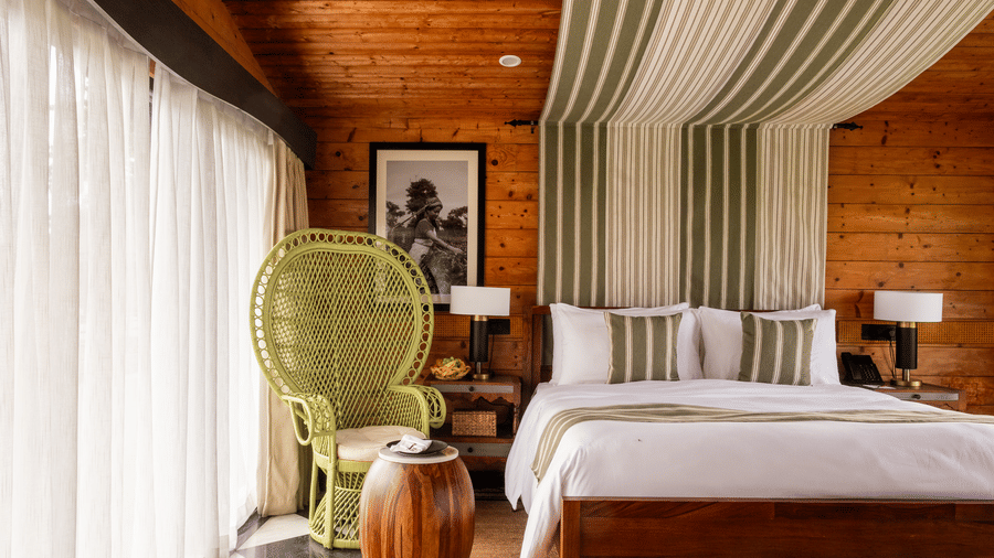 A complete view of a bedroom at Rhino & River Wildlife Retreat & Spa, Pobitora, showcasing a white bed, striped canopy, wicker chair, and large windows with white curtains.