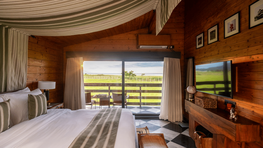 A luxury suite at Rhino & River Wildlife Retreat & Spa, Pobitora, featuring a large bed, striped ceiling canopy, and sliding glass doors leading to a balcony.
