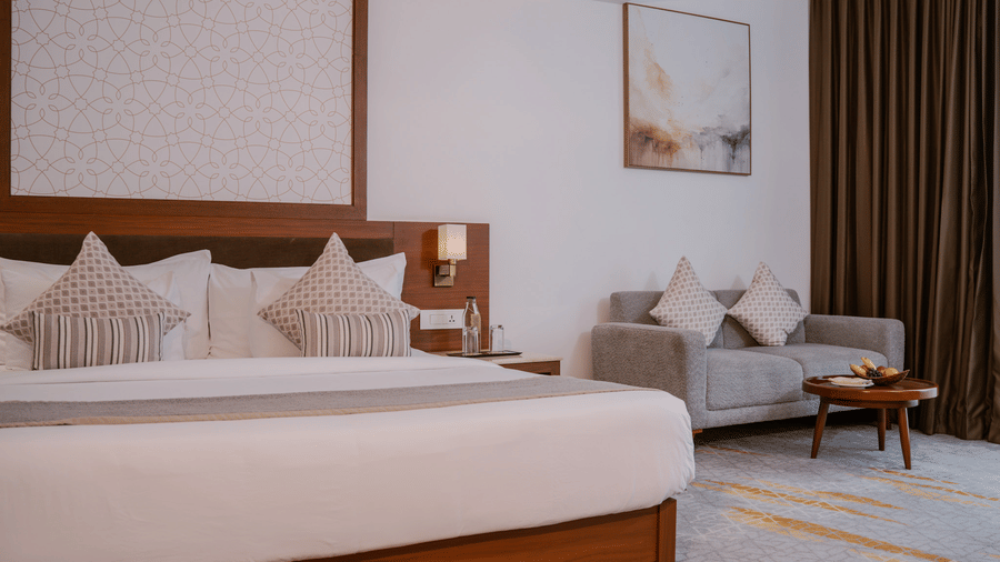A bedroom features a wooden king size bed with patterned cushions next to a grey 2 seater sofa and a round coffee table at Tiara by MPS.