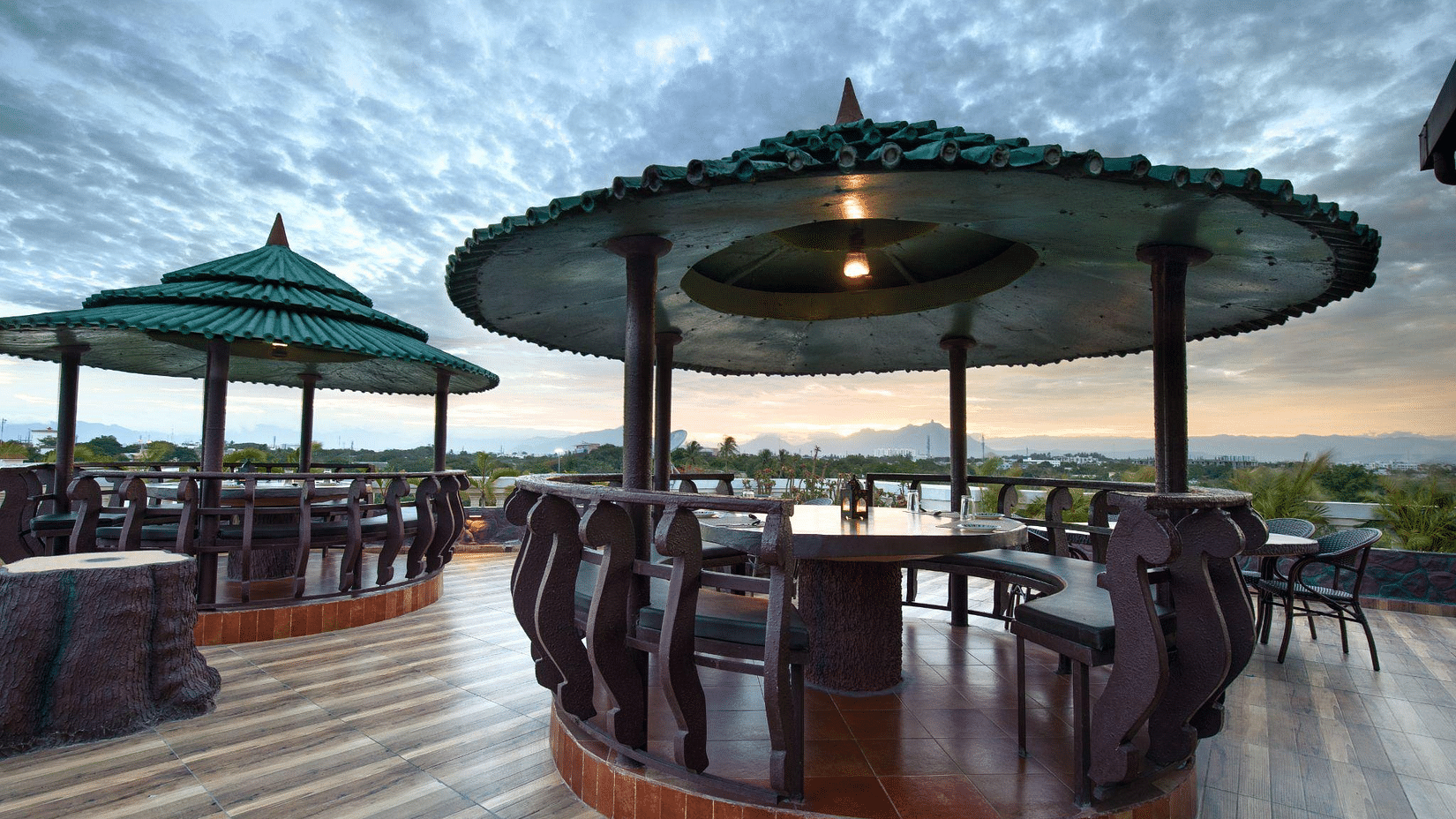 Outdoor dining area with several covered gazebos featuring tables and chairs on a wooden deck, set against a dramatic, cloudy sky - Jenneys Residency, Coimbatore