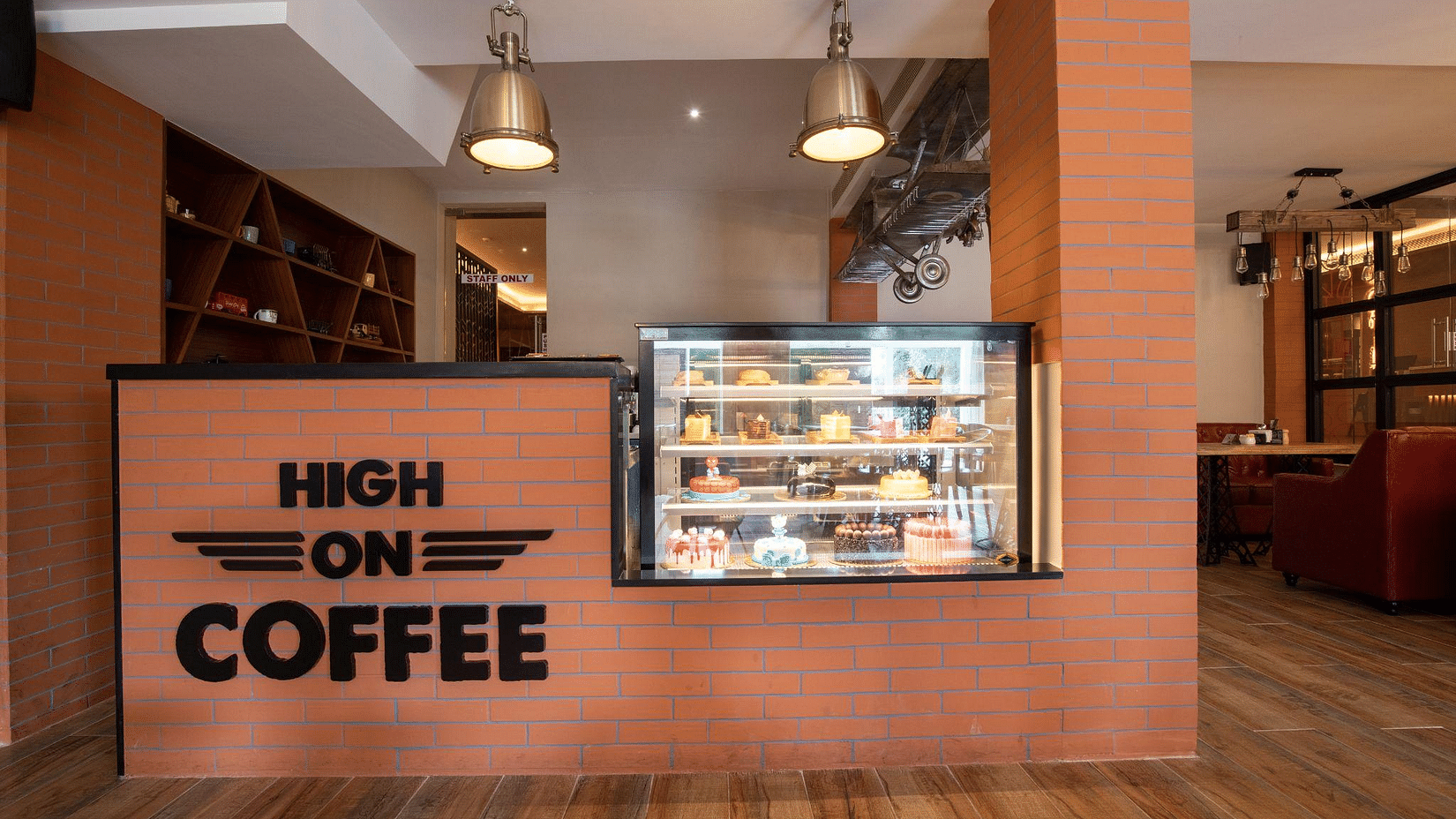 Cafe counter with High On Coffee signage, a display fridge filled with pastries, pendant lighting, and a glimpse of the seating area - Jenneys Residency, Coimbatore