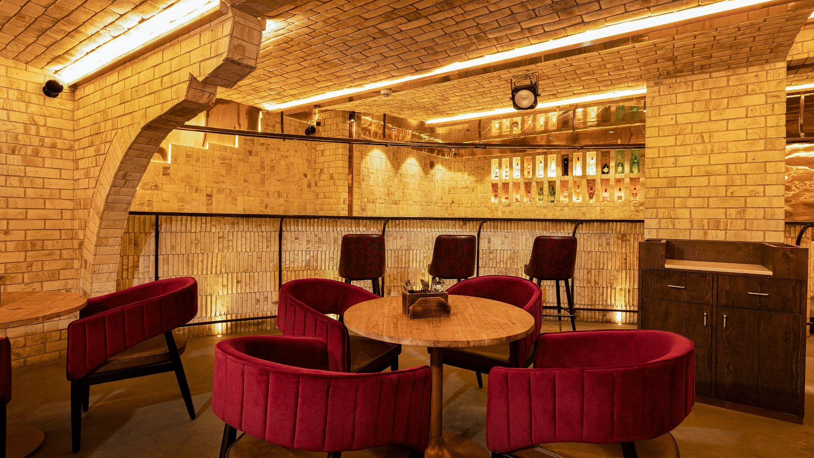 Cosy bar area with exposed brickwork, warm lighting, a central round table with plush red chairs, and bar stools along a counter - Jenneys Residency, Coimbatore