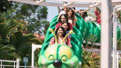 Group of smiling people riding a green caterpillar roller coaster at an amusement park.
