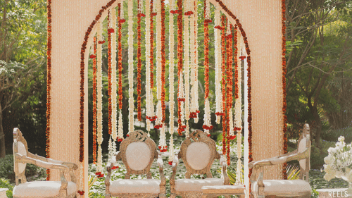 Ornate wedding mandap with floral backdrop and traditional chairs set in a garden.