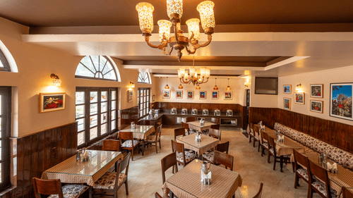 The dining area of Buransh Restaurant with set tables, wooden chairs, chandeliers, and framed artwork on the walls at The Pavilion, Nainital