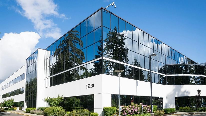 A modern office building with reflective glass windows and white panels stands under a bright blue sky with clouds.