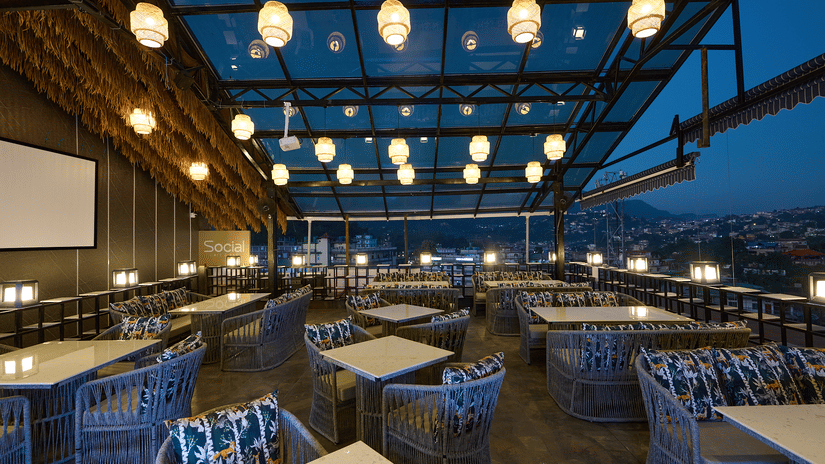 An outdoor or rooftop restaurant with tables and chairs under a glass ceiling, illuminated by hanging glass globe lights, with a nighttime view of a city or landscape in the background | Voyage Eastin Suites