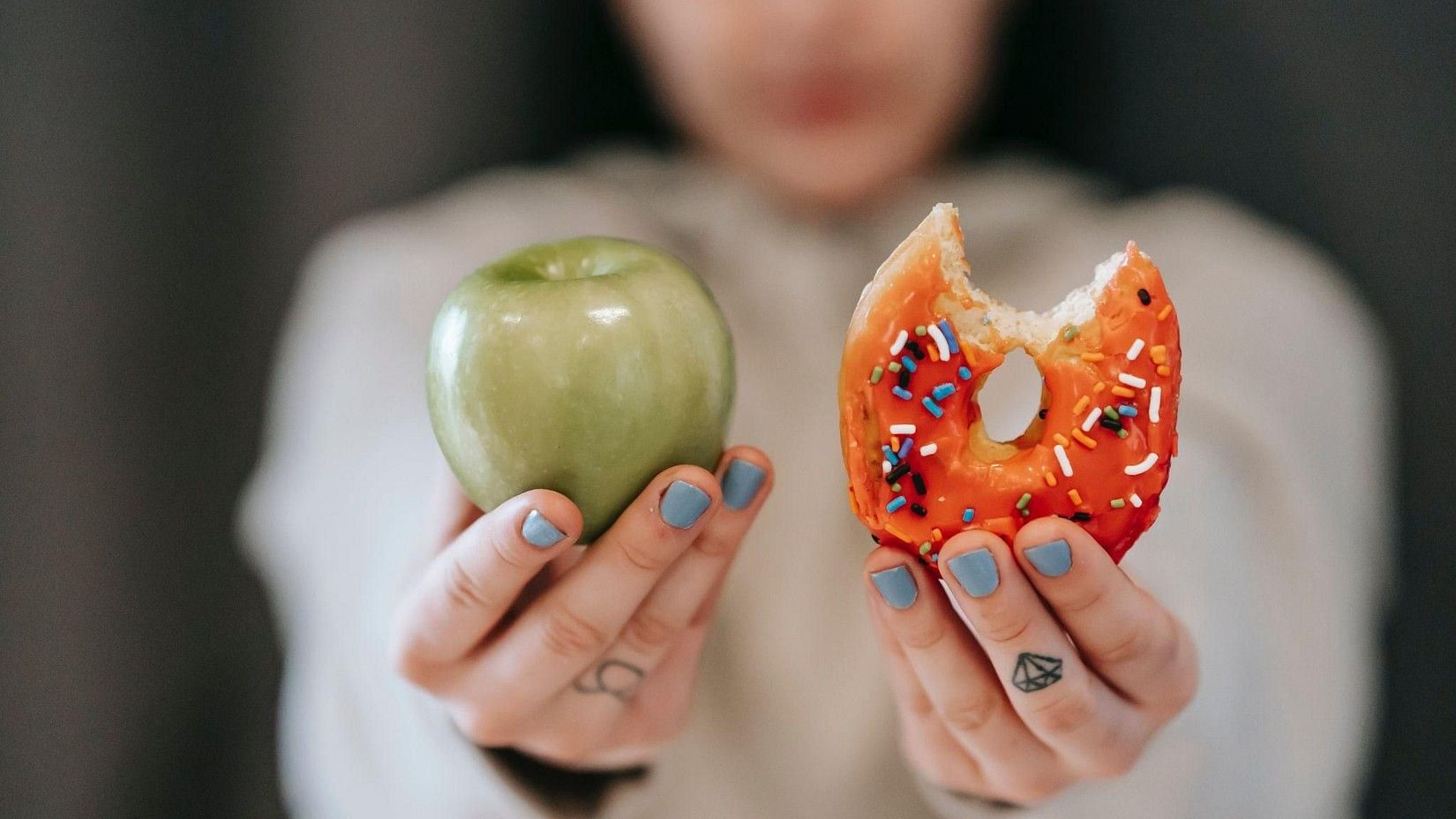 Hands hold a green apple in one hand and a half-eaten, sprinkles-covered donut in the other