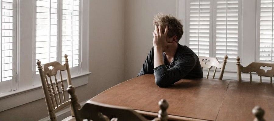 Person sitting alone at a wooden dining table in a sunlit room, resting their head on their hand, appearing contemplative
