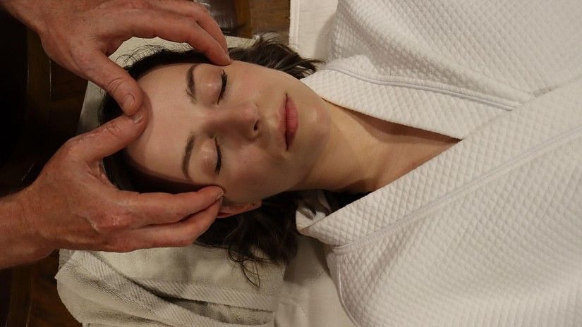 A woman in a white robe lying down with closed eyes receiving facial massage