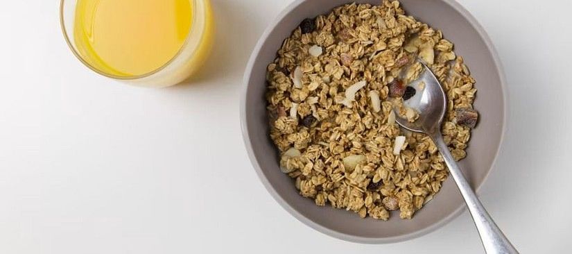 a glass of orange juice and a bowl of granola with a spoon, on a white surface.