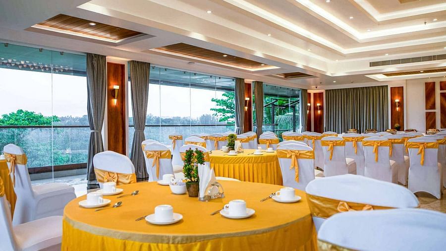 A bright and airy event hall at Mansha Palace is set up with round tables draped in yellow and white chairs with yellow sashes, offering a pleasant view through large windows.