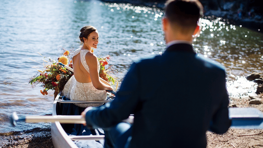 Bride seated in a boat holding a bouquet while facing the lake with the groom standing in the foreground at Kit Carson Lodge.