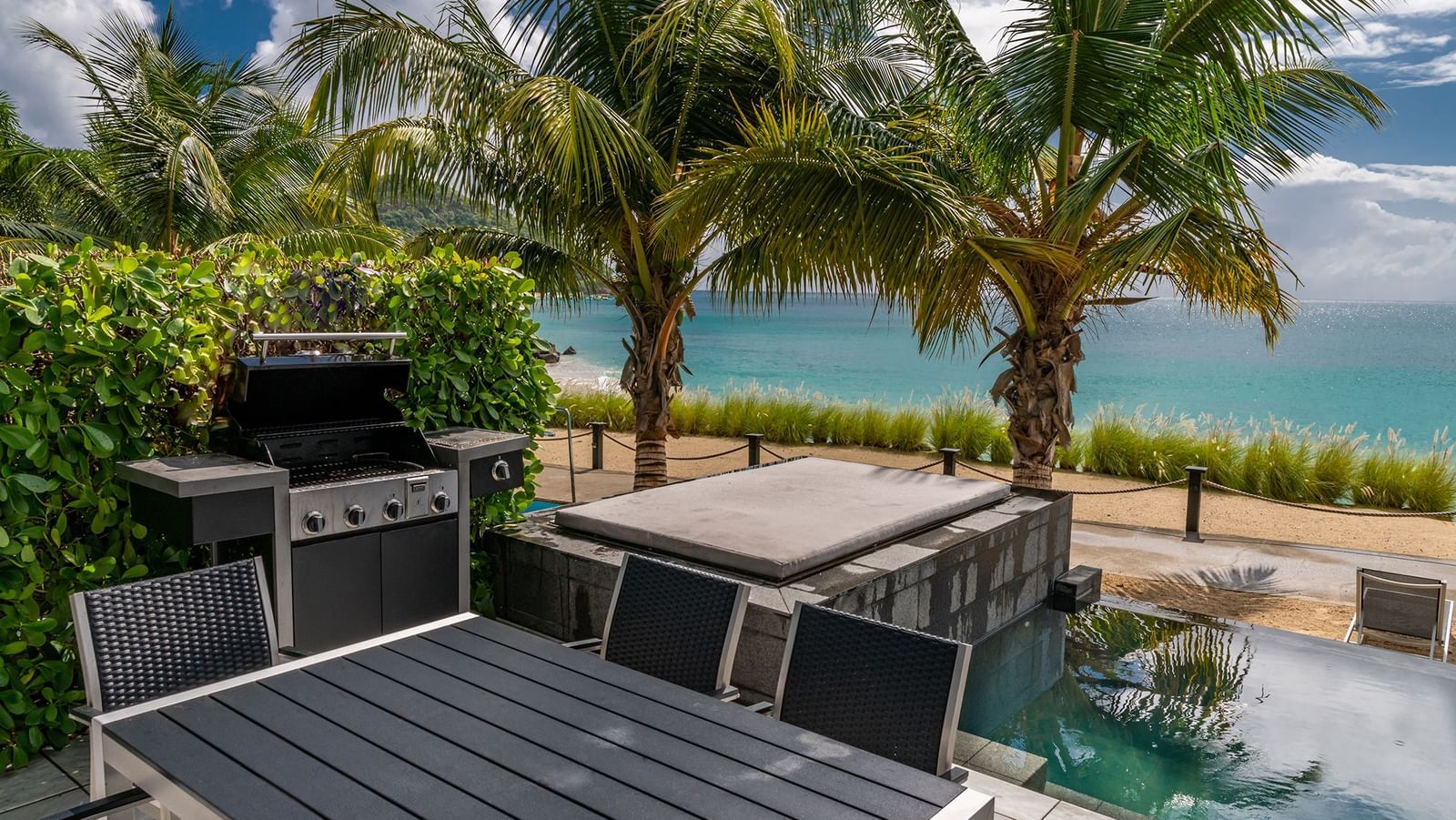 An outdoor dining area with a barbecue grill and hot tub overlooks the ocean, framed by palm trees and lush greenery. 