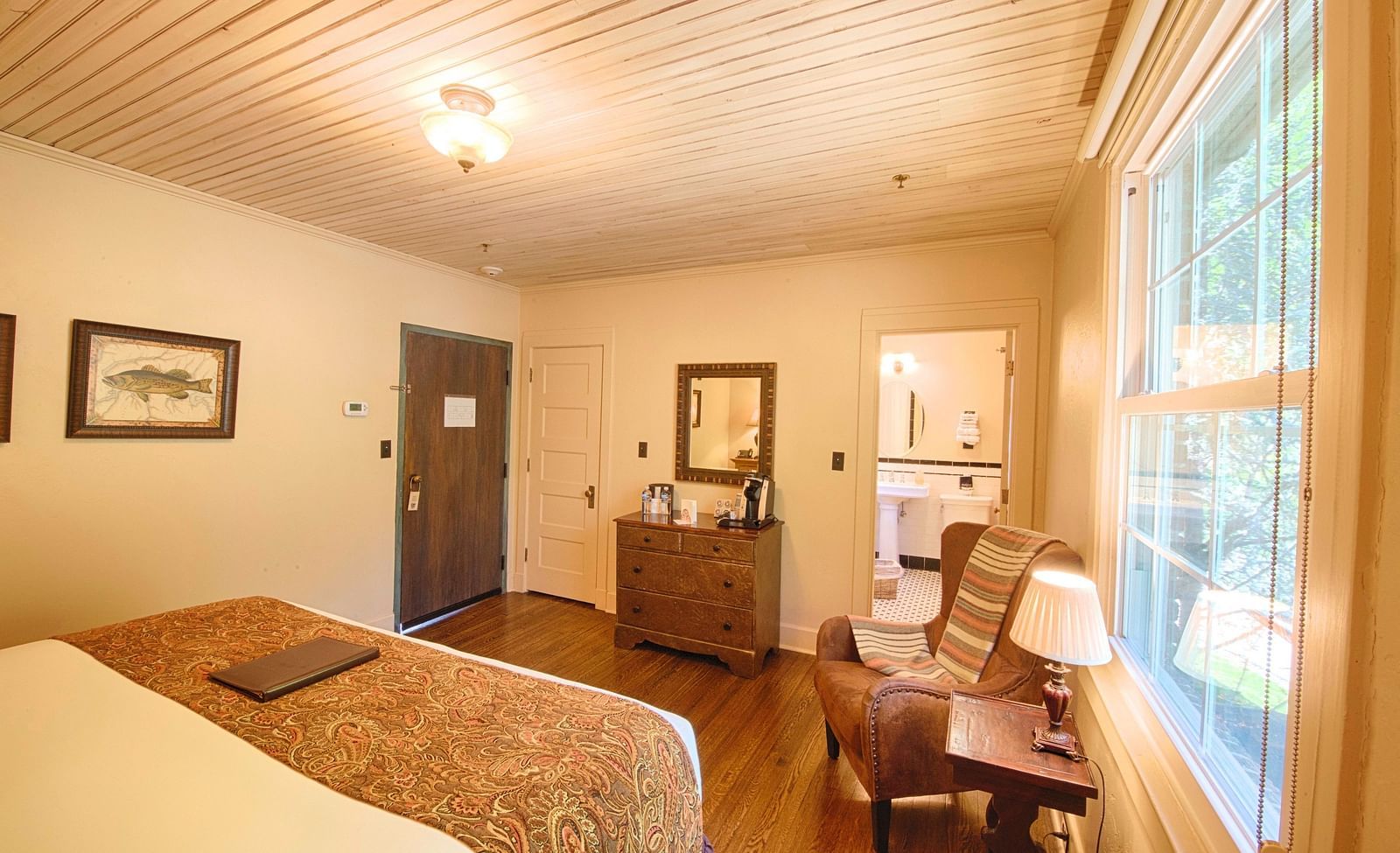 a cozy bedroom with a cushioned rustic armchair along with a small side table with a table lamp and a double bed in the room - Historic Tapoco Lodge